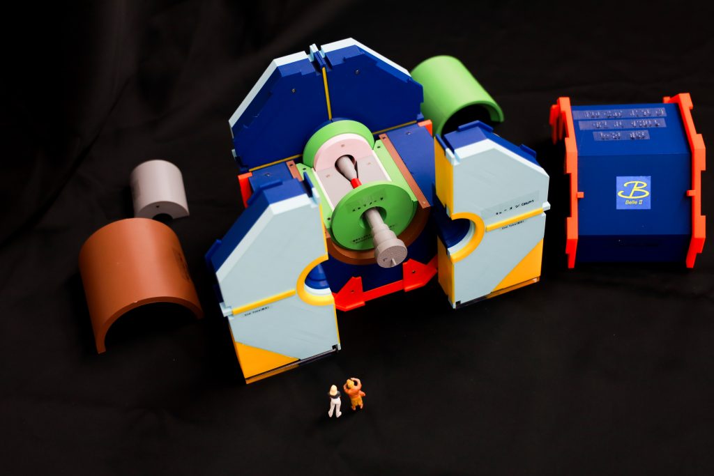 KEK で稼働する巨大な素粒子測定器（約8m）を1/40 スケールで再現した模型の写真。分解できることがわかるように、一部は分解された状態で置かれている。測定器の模型とともに人形も置いてあり、スケール感がわかるようになっている。