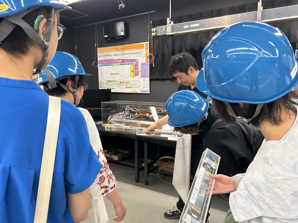 写真中央で研究者が、行なっている実験について説明している。研究者を取り囲むように筑波技術大学の学生と教員が立っている。右手前の見学者が実験装置の一部を手にしている。